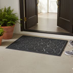 Black doormat with geometric pattern on a light-colored floor, with open doors and potted plants in the background.