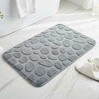 Gray textured bath mat on a white surface with white slippers and a plant.